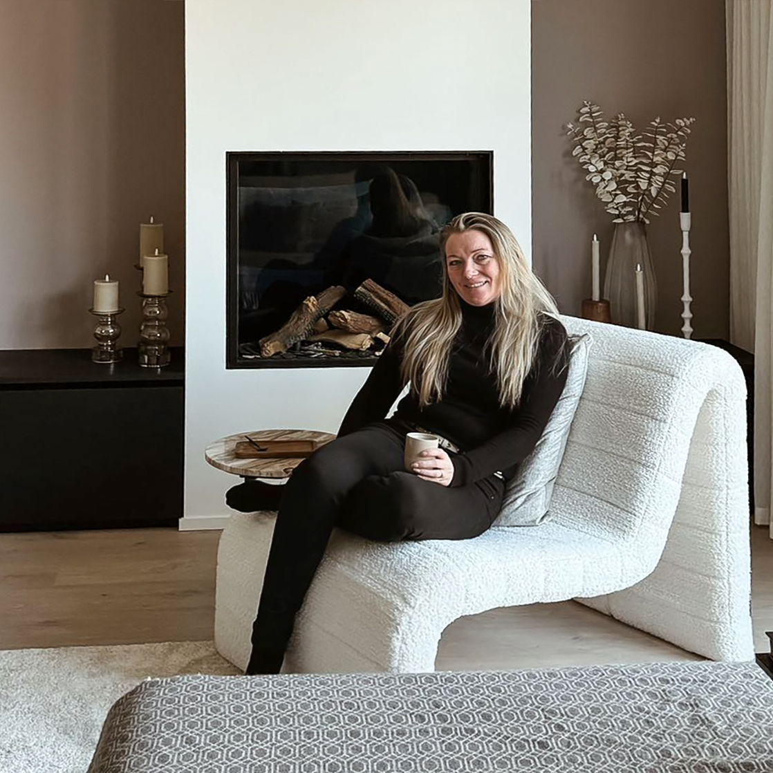 Vrouw zittend op witte fauteuil met kopje in haar hand in sfeervol ingerichte woonkamer met open haard. Dat is Jenny en haar stijl
