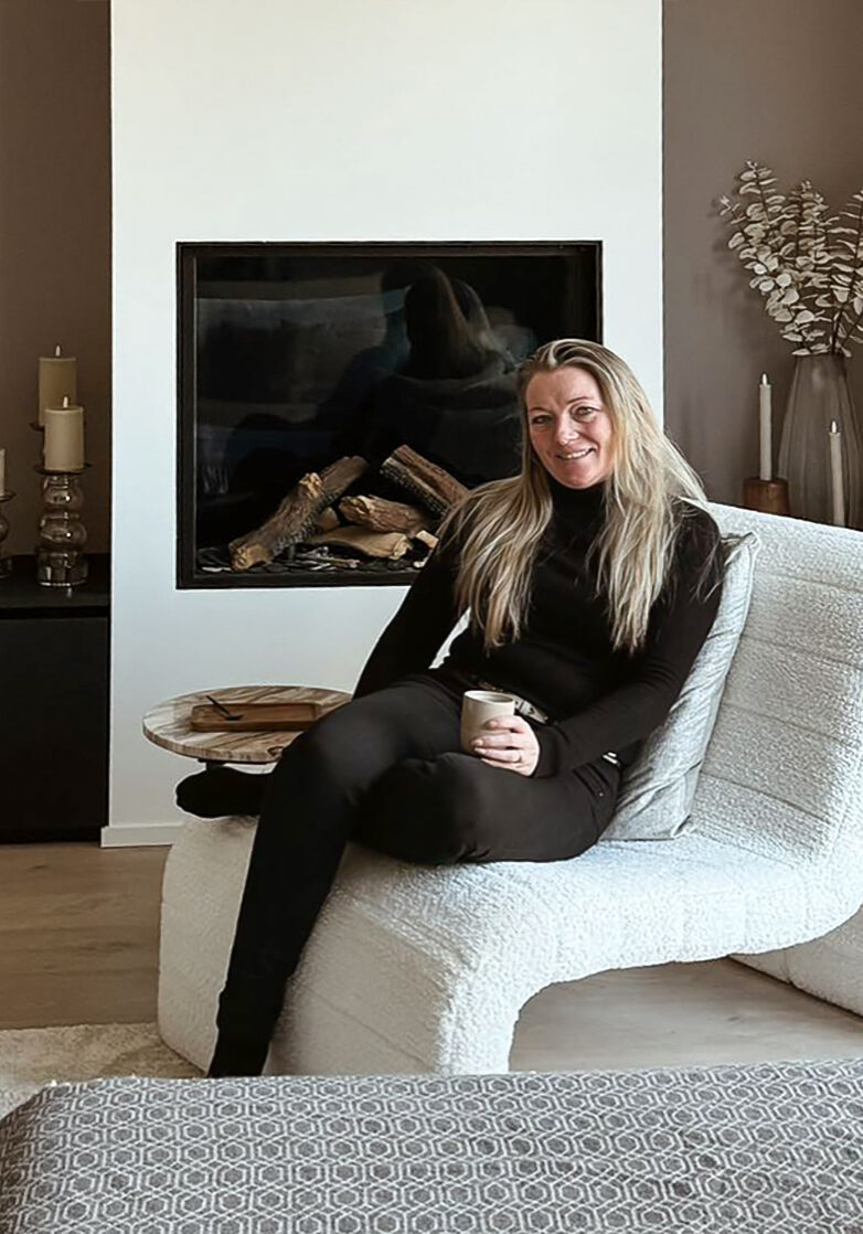 Vrouw zittend op witte fauteuil met kopje in haar hand in sfeervol ingerichte woonkamer met open haard. Dat is Jenny en haar stijl