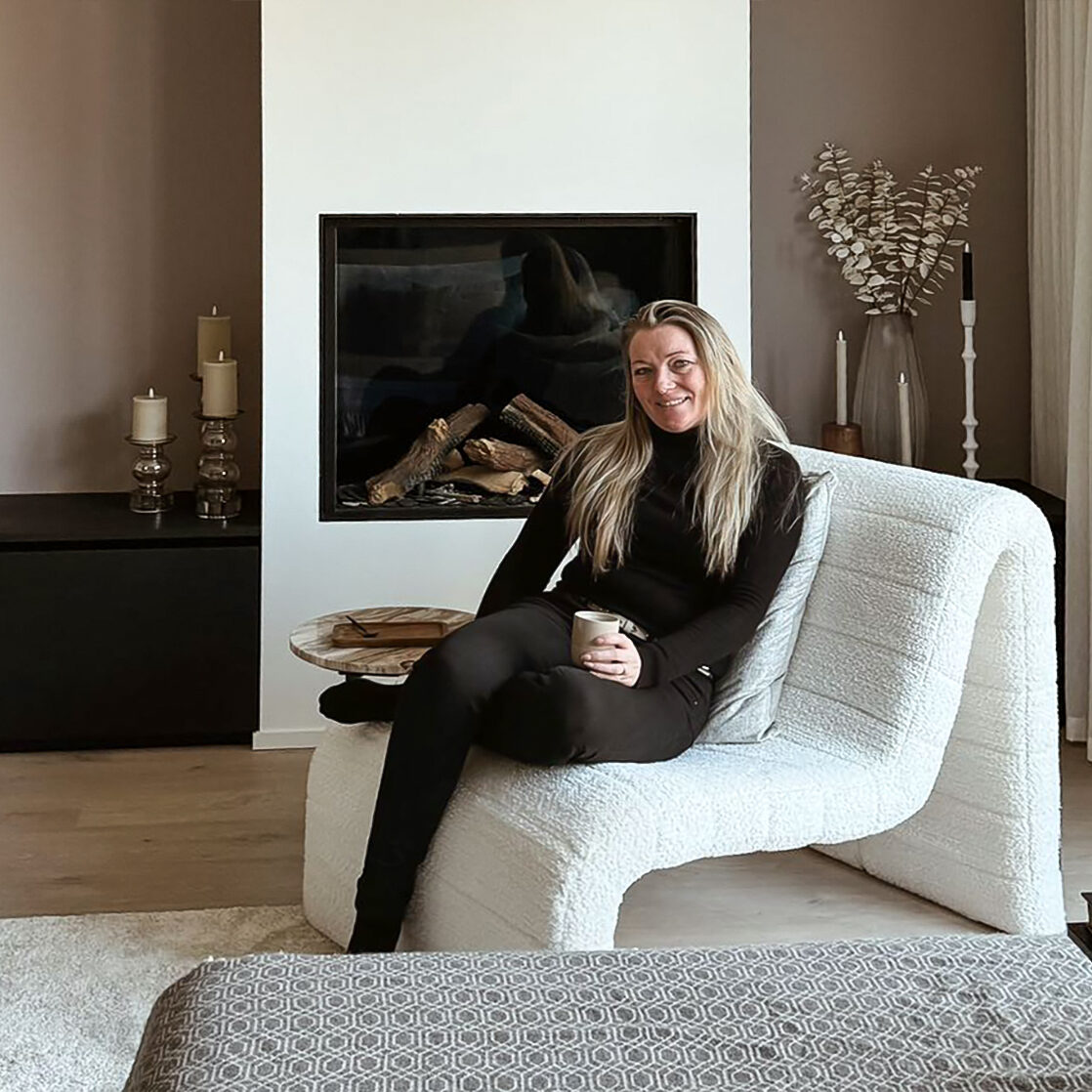 Vrouw zittend op witte fauteuil met kopje in haar hand in sfeervol ingerichte woonkamer met open haard. Dat is Jenny en haar stijl